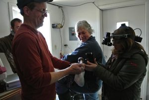 A man in a baseball cap holds a bird while Dr. Gillian McLellan performs and eye exam. Colleagues in the background are observing